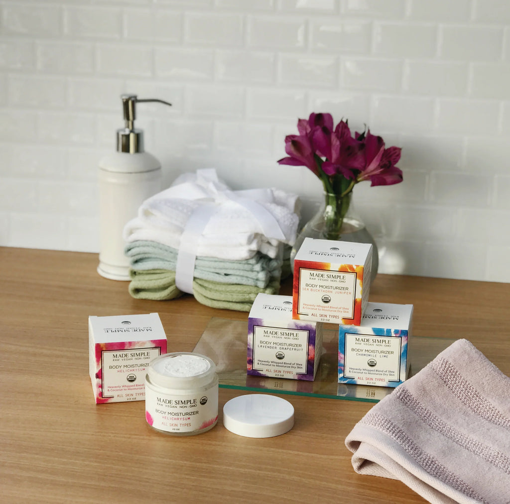 Natural body moisturizer jars and boxes on a wooden bathroom counter with towels, soap dispenser, and vase of purple flowers.