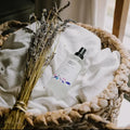 Lavender linen mist spray bottle with dried lavender in a woven basket on white fabric