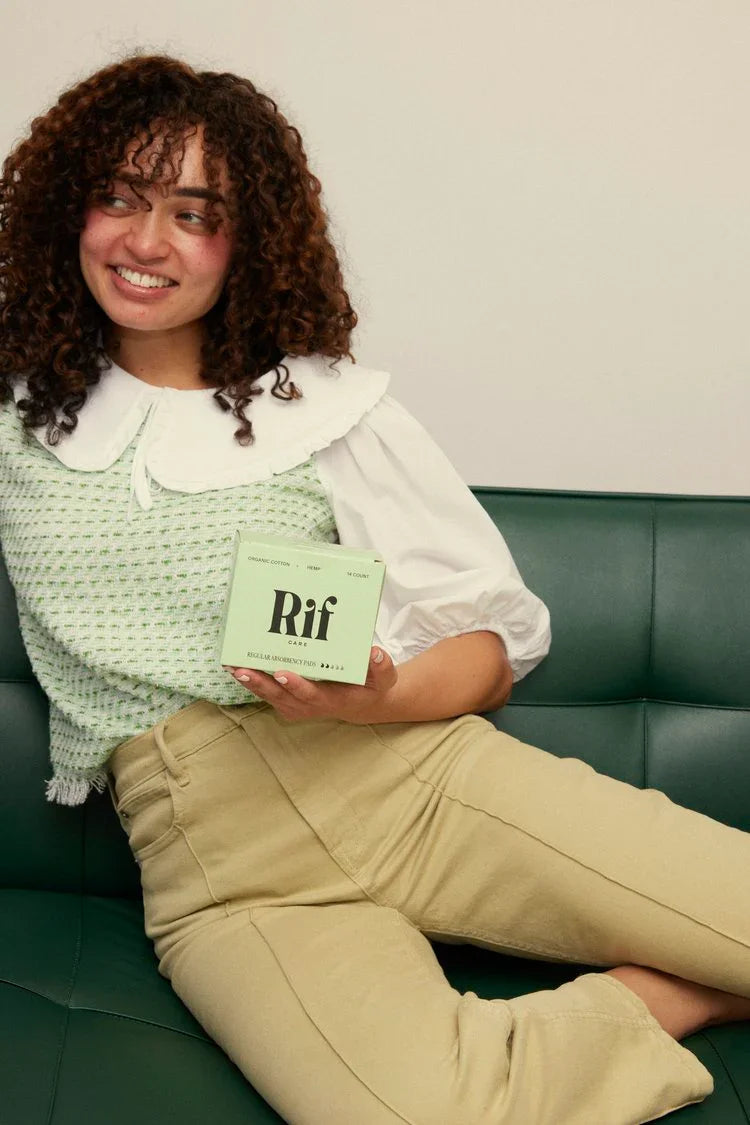 Smiling woman sitting on green couch holding a box of organic cotton hemp period pads